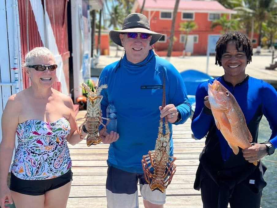 Three people smile while holding freshly caught lobsters and a fish on a sunny dock.