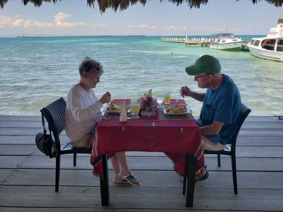 An older couple enjoys a meal at a seaside table with a view of the water and boats.