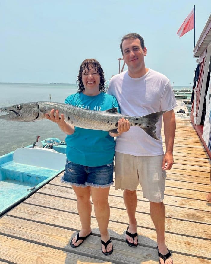 A woman and a man pose together on a dock, holding a large fish against a backdrop of water and sky.
