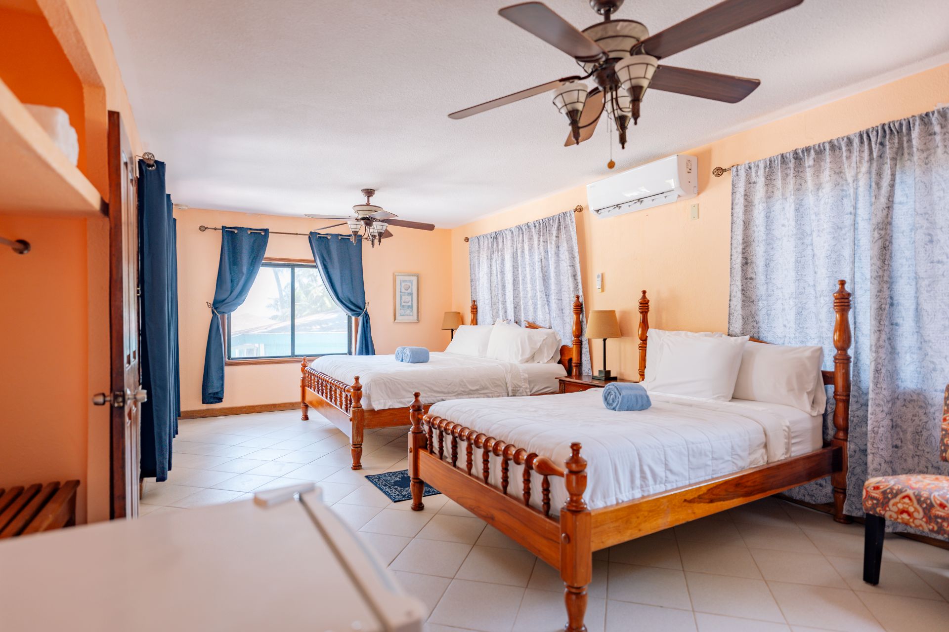 Brightly lit bedroom featuring two wooden beds, blue curtains, and a window with a view.