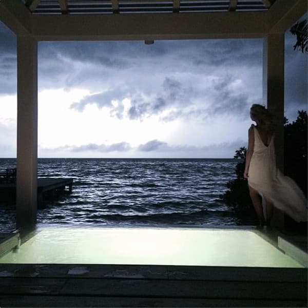 A woman in a flowing dress stands by a shallow pool, gazing out at a stormy ocean under a moody sky.