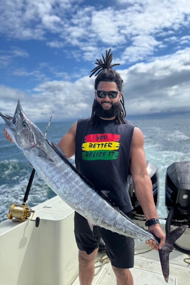 A person with dreadlocks and sunglasses holds a large fish while standing on a boat, with a cloudy sky in the background.