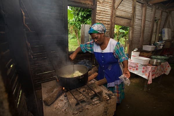 A woman stirs a large pot on a wooden stove inside a rustic kitchen.
