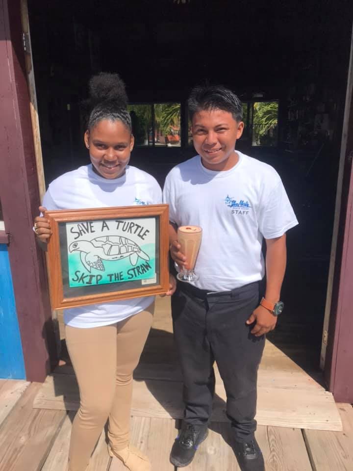 Two staff members hold a framed sign promoting turtle conservation and sustainable practices, with one holding a drink.