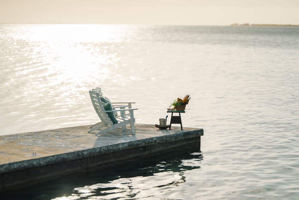 A white adirondack chair and a small table with flowers sit on a dock by the water.