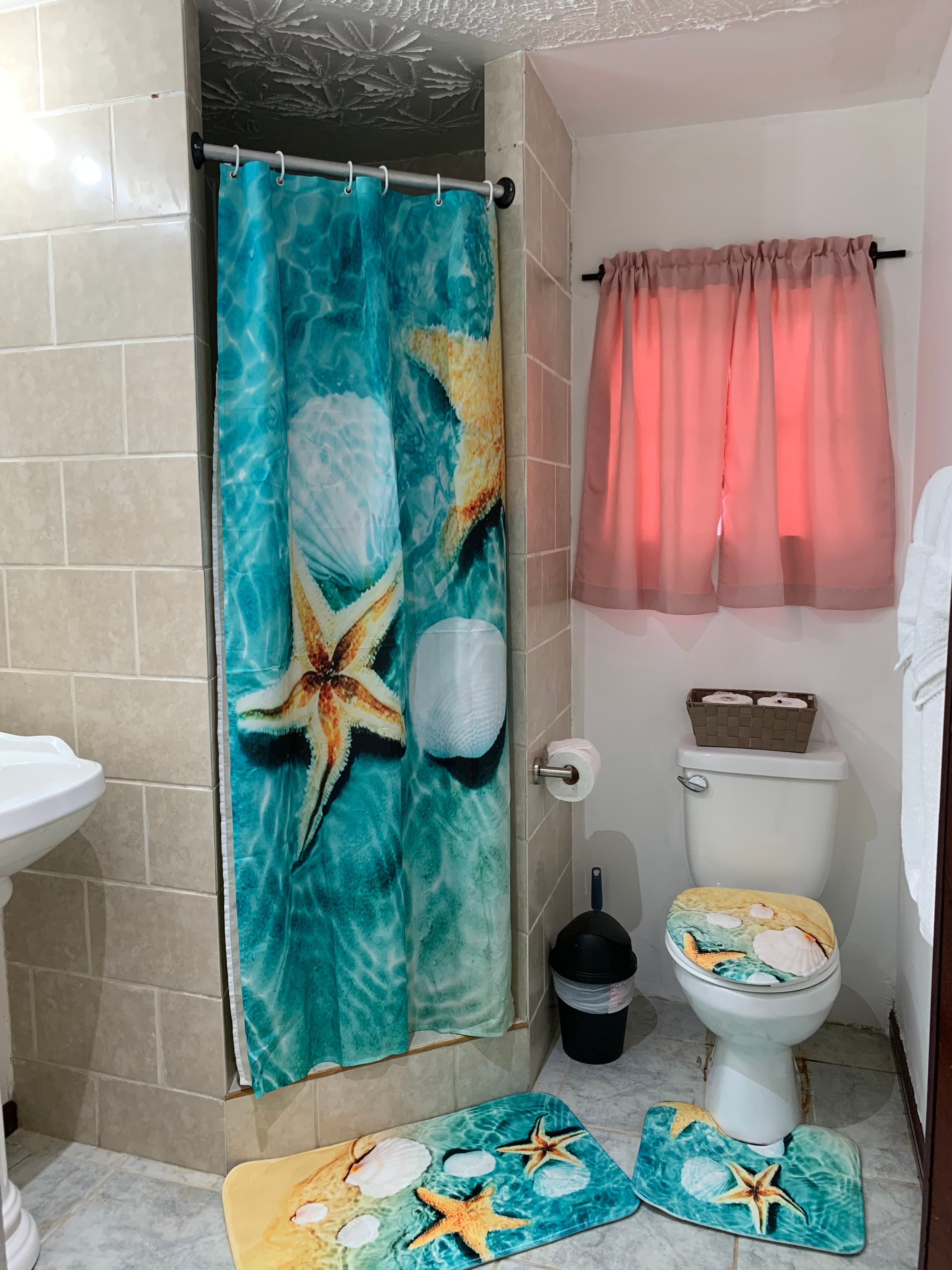 A beach-themed bathroom featuring a starfish shower curtain, matching bath mats, and a window with pink curtains.
