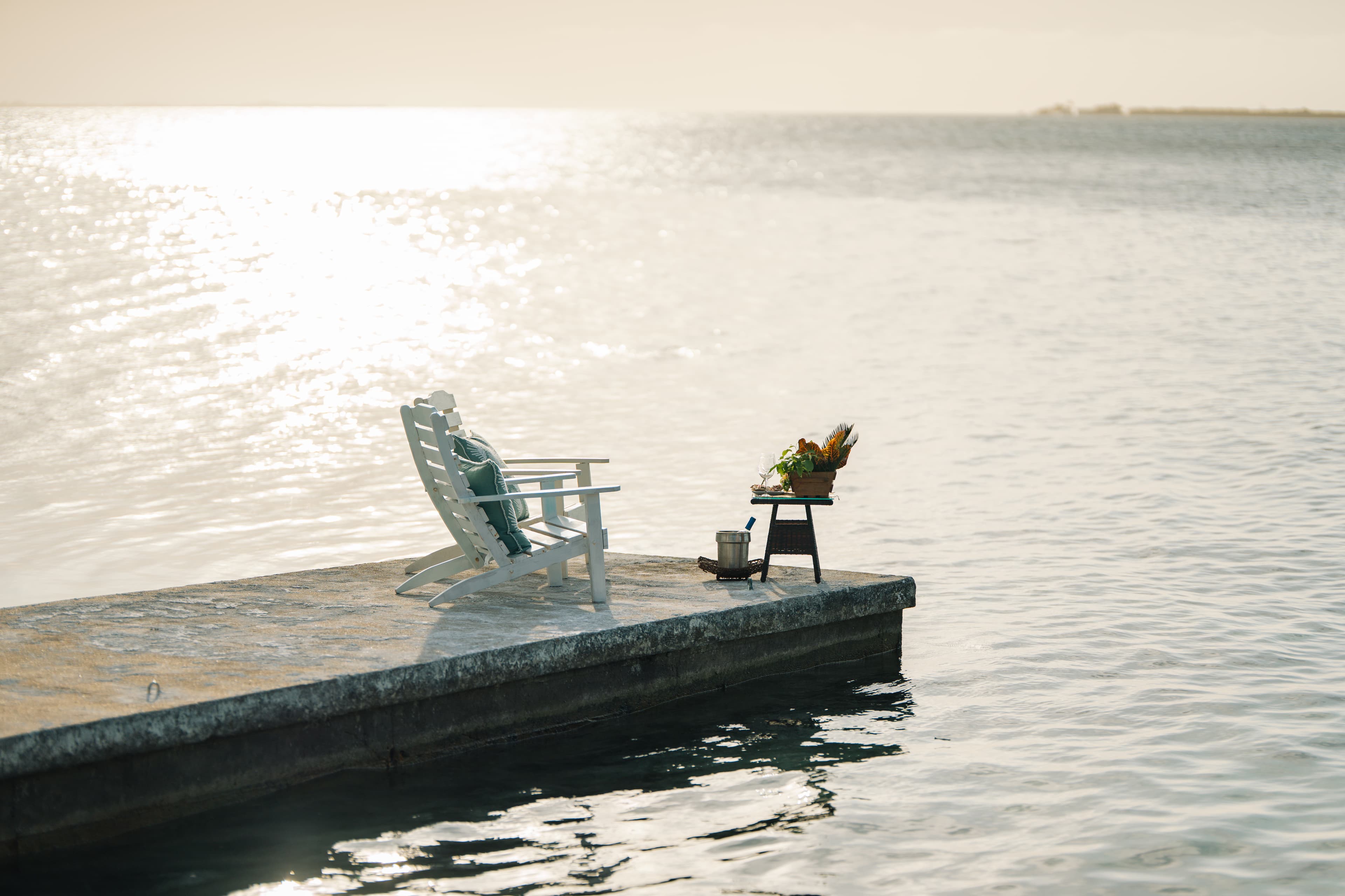 A white adirondack chair and a small table with plants sit on a dock overlooking water.