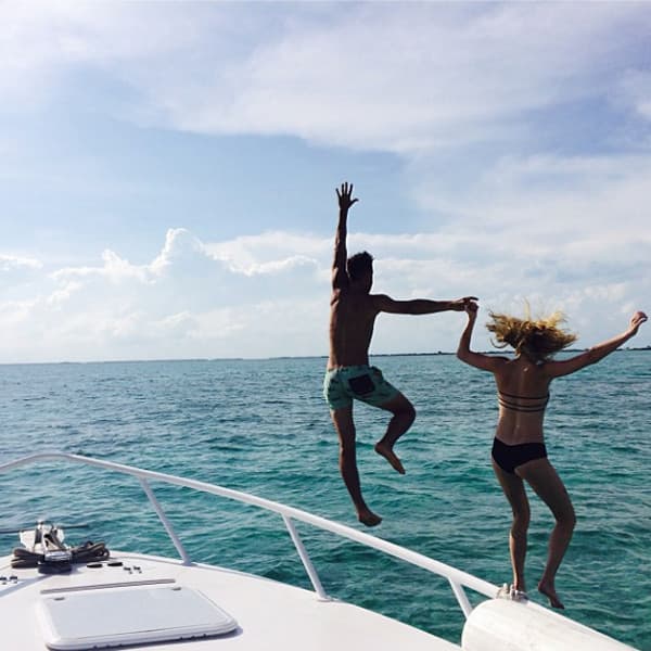 A man and woman jump off the bow of a boat into clear blue water under a sunny sky.