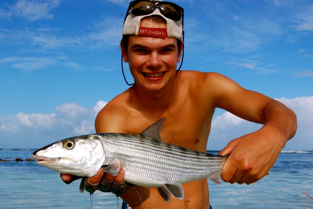 A young man smiles while proudly holding a large fish against a sunny beach backdrop.