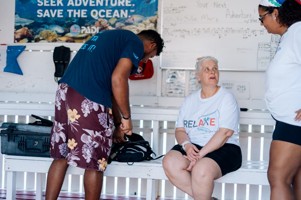 Three people engage in conversation in a dive shop, with one preparing gear.