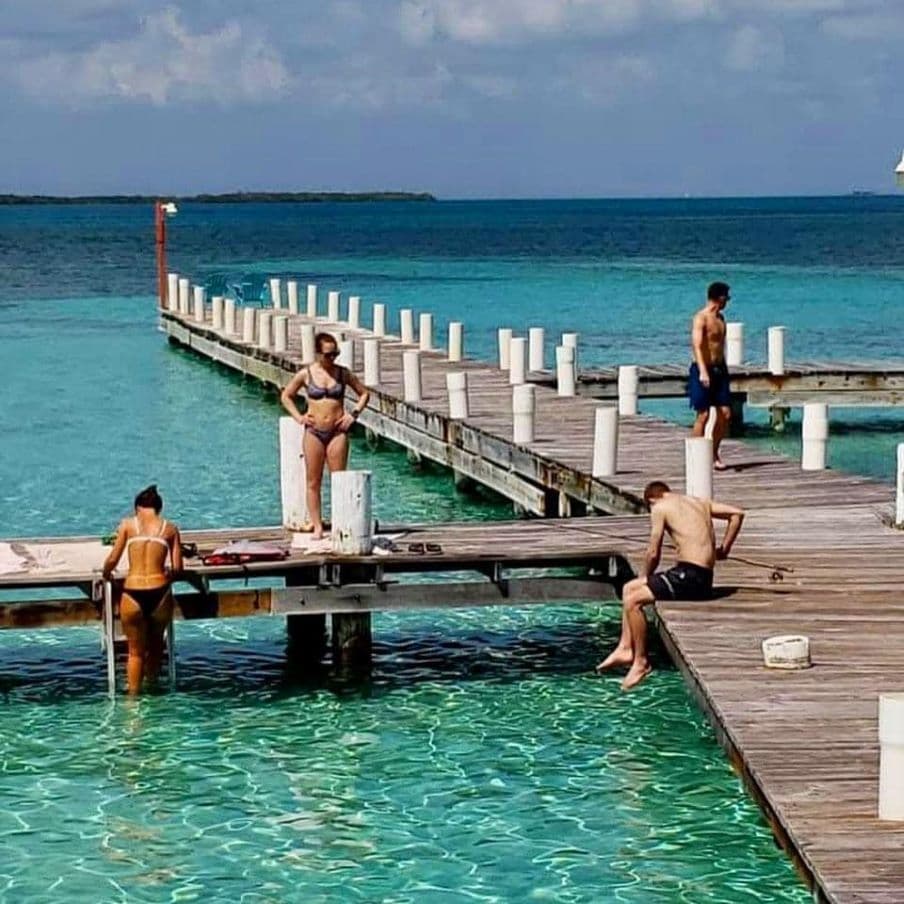 Four people enjoy a sunny day by a wooden pier extending into clear turquoise water.