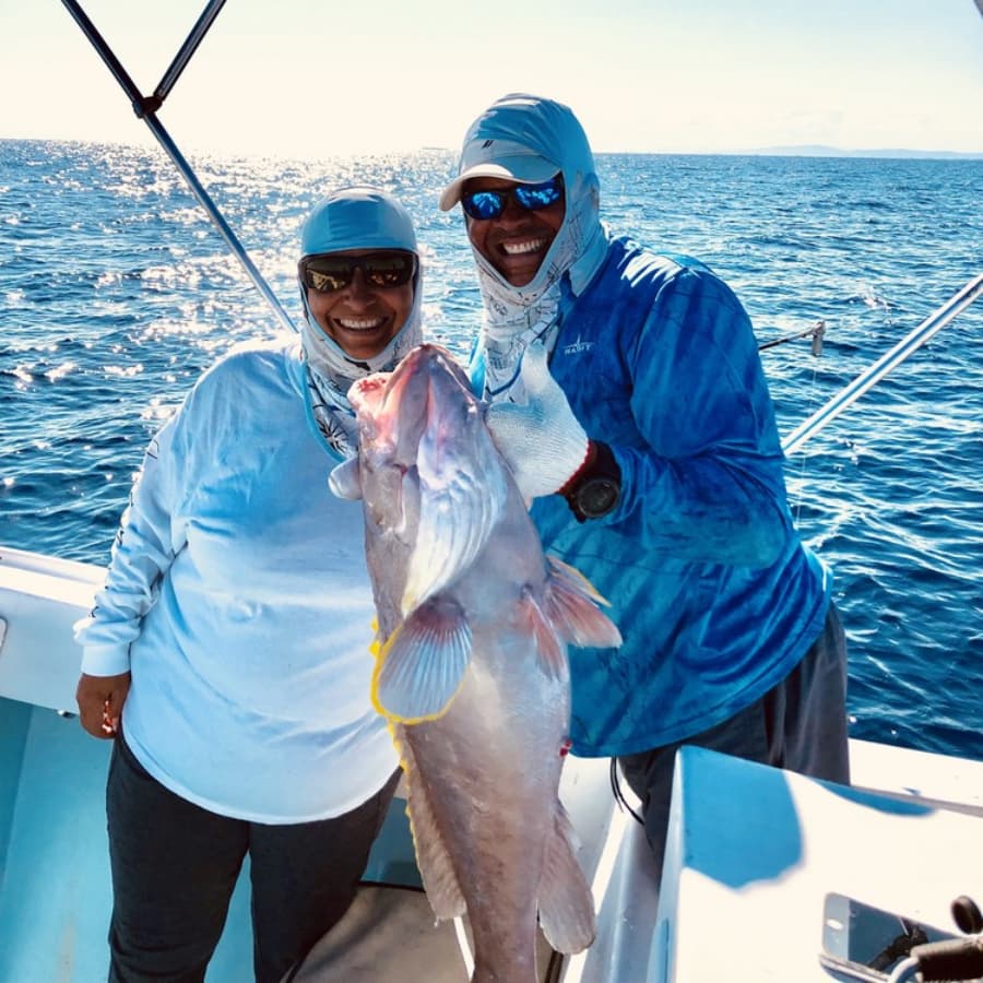 Two smiling people on a boat hold up a large fish against a shimmering ocean backdrop.