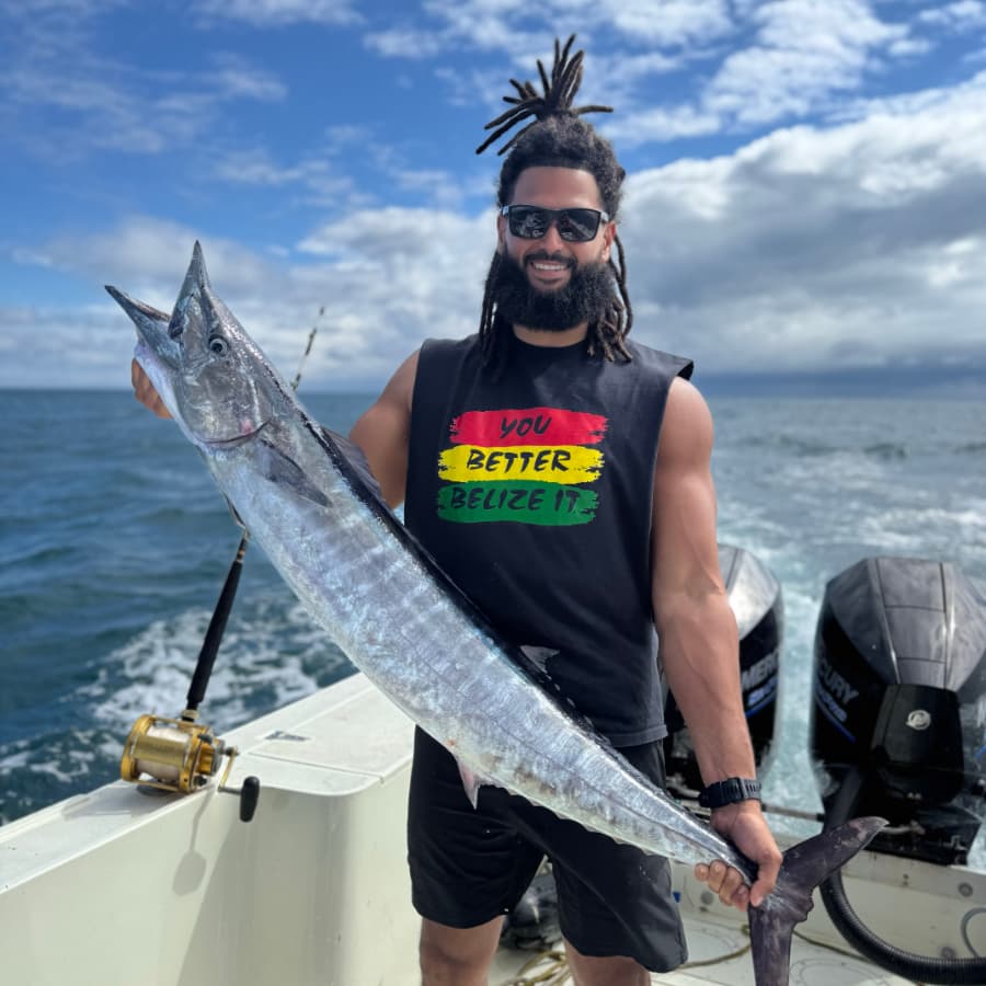 A smiling man with dreadlocks holds a large fish while on a boat under a blue sky.