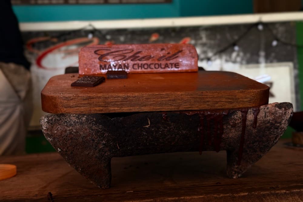 A wooden block with "Cheri" and "Mayan Chocolate" engraved on it sits atop a rustic stone base, with melted chocolate dripping off the edges.