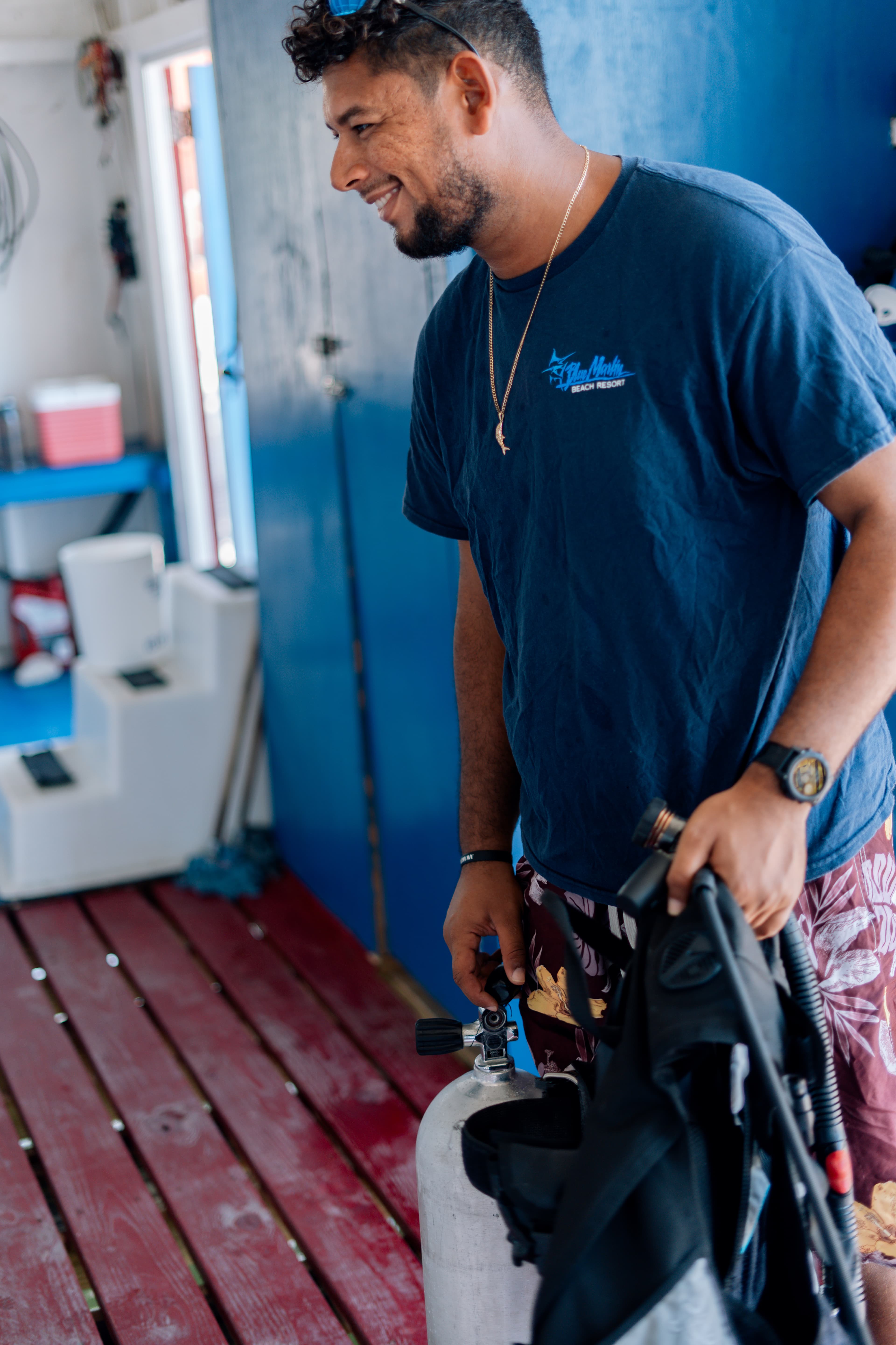 A smiling man holds a scuba tank and diving gear inside a diving shop.