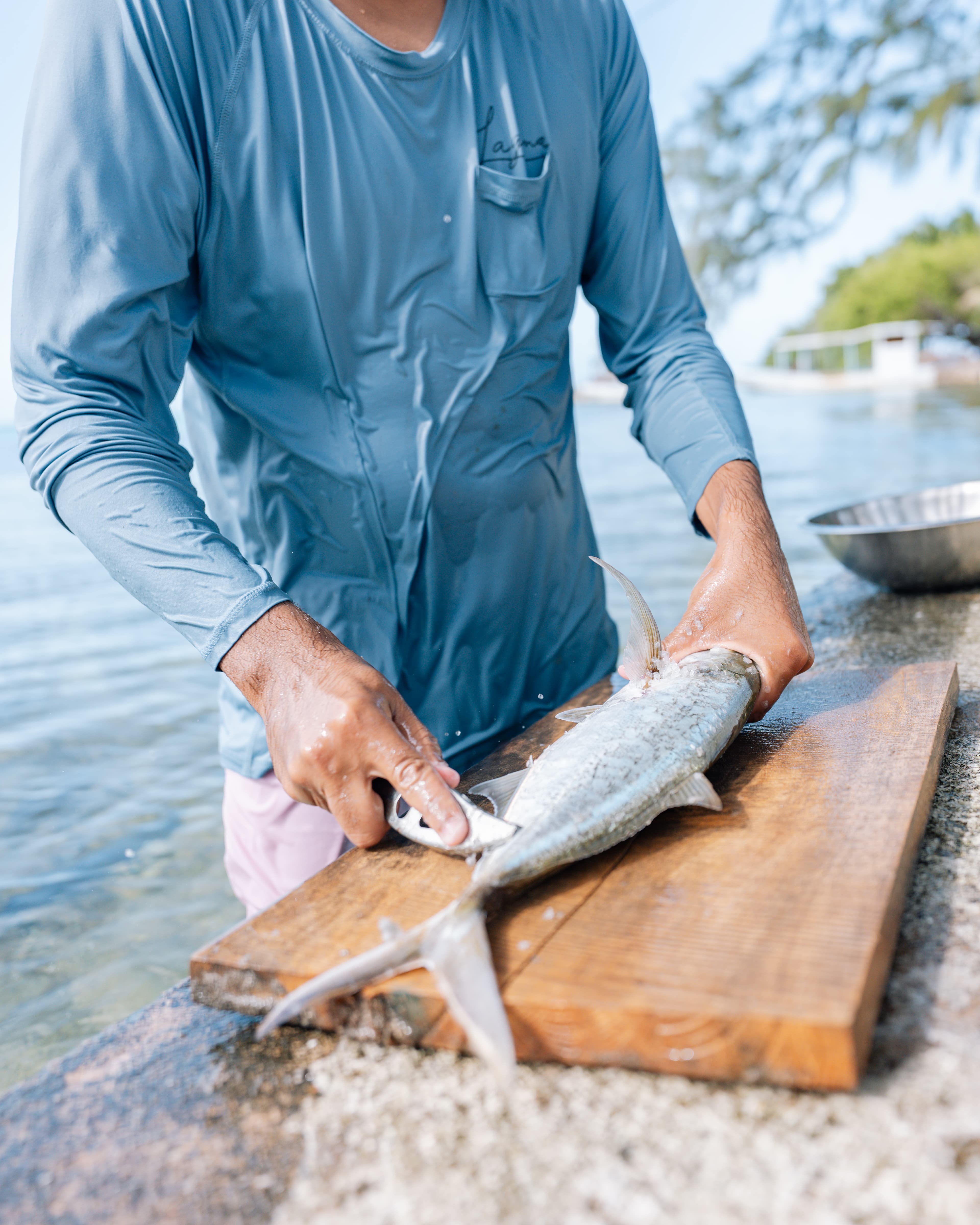 A person in a blue shirt is filleting a fish on a wooden cutting board by the water.