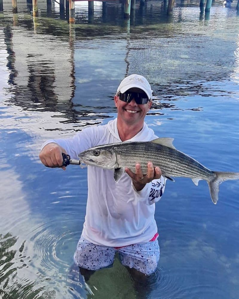 A man stands in shallow water, holding a fish and smiling.
