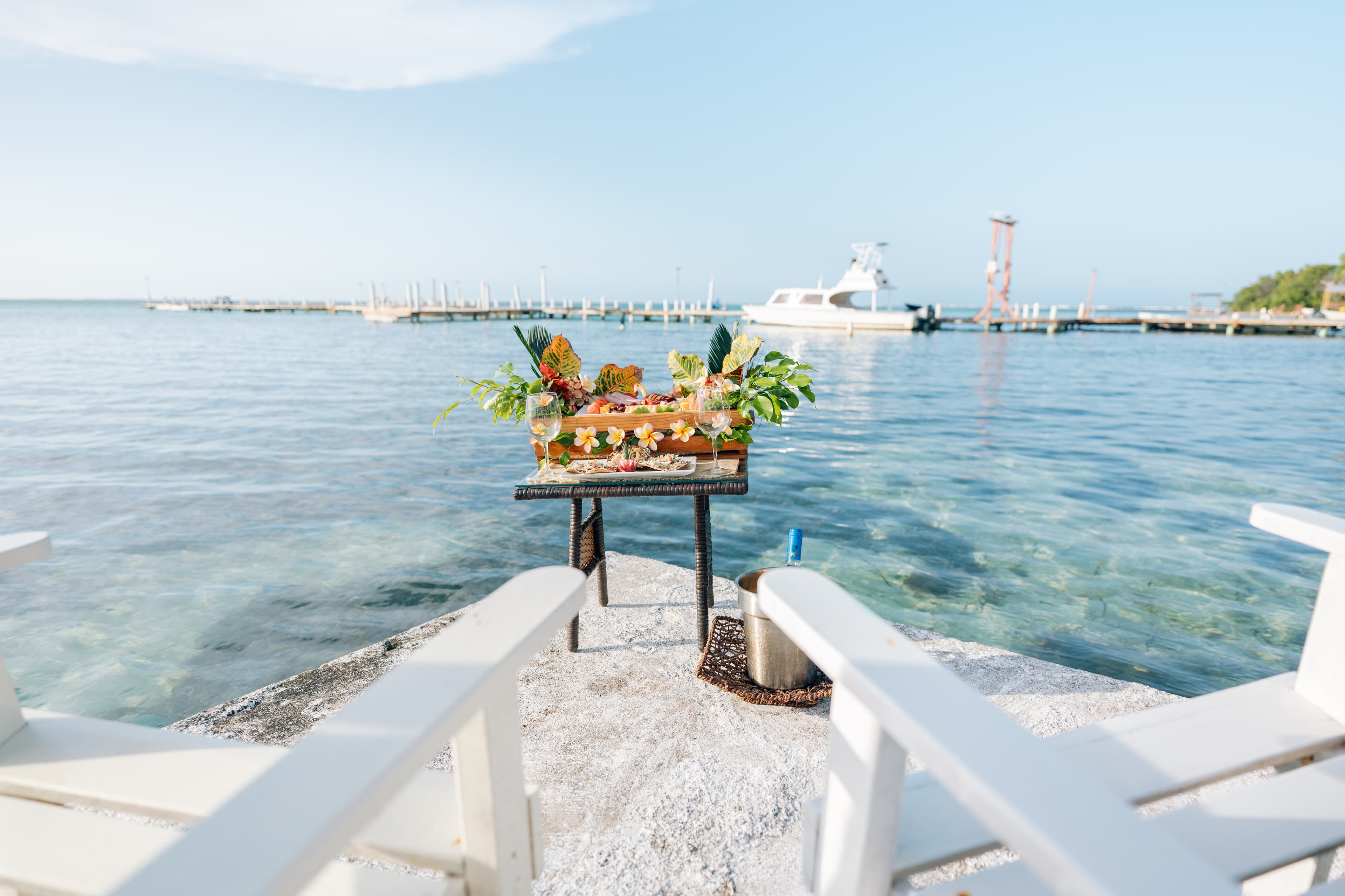A vibrant display of tropical fruits and drinks is set on a table by the calm, clear water, with a pier and boat in the background.