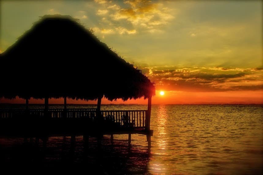 Silhouetted thatched roof on a dock at sunset over calm waters.
