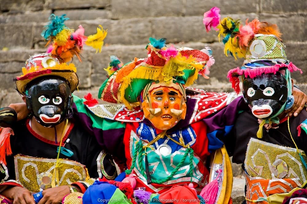 Three individuals wearing colorful traditional attire and masks, adorned with feathers, pose together.