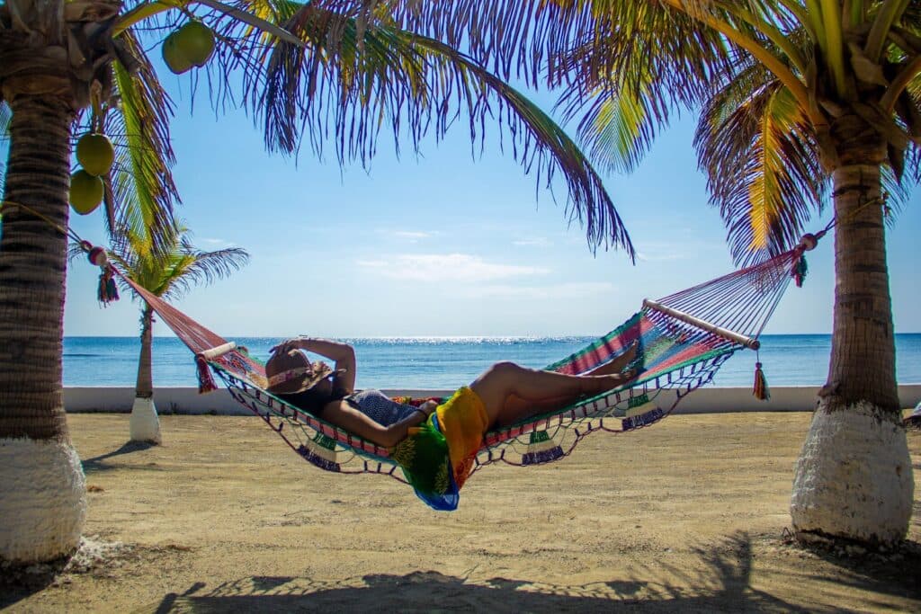 A person relaxes in a colorful hammock between palm trees on a sunny beach.