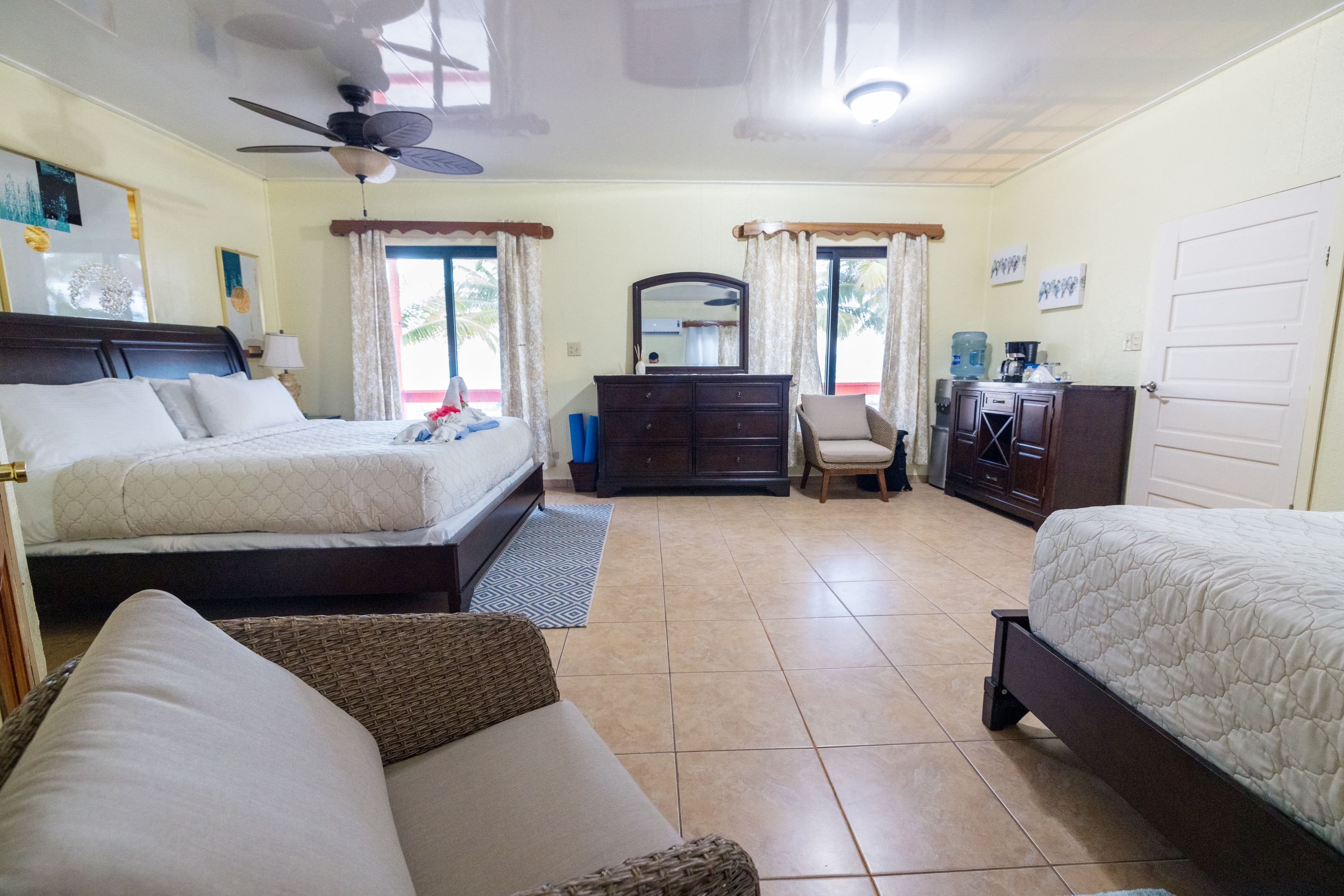 A spacious hotel room featuring two beds, a dresser, and a sitting area with natural light from large windows.