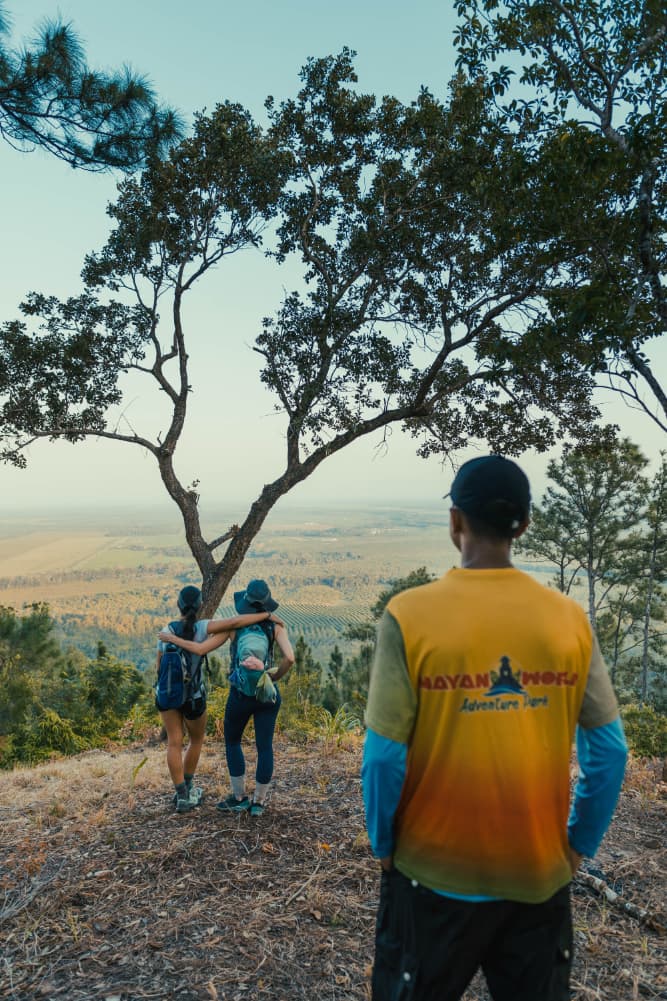 Three hikers enjoy a scenic view from a hillside, with two women embracing and a man standing nearby.