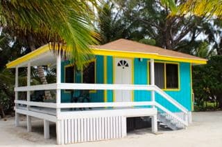 A blue cottage with yellow and white trim on a sandy island surrounded by palm trees