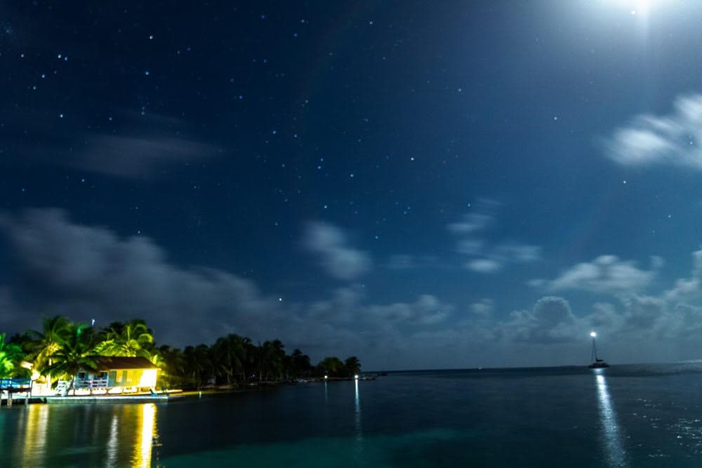 A beautiful midnight sky over the ocean and the private island of South Water Caye with the sky dotted with lots of stars, and the island buildings lit up, along with a lit sailboat on the water.