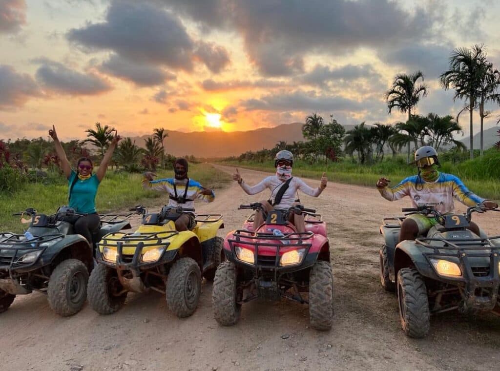 4 smiling people with thumbs up on ATVs on a dirt road through the jungle with the sun setting behind them.
