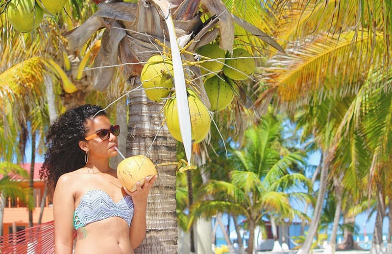 A woman at a tropical resort surrounded by palm trees drinking coconut water from a coconut