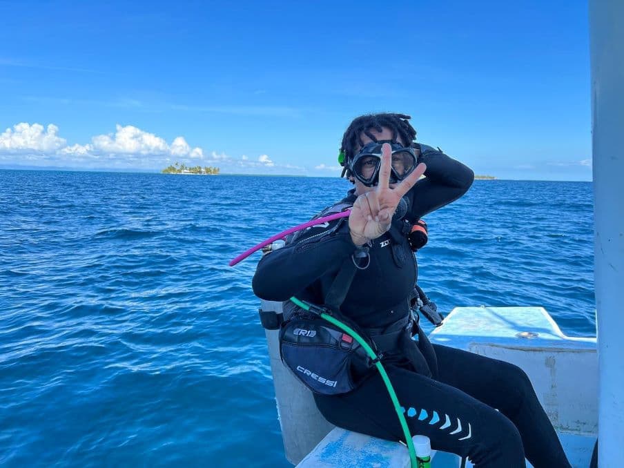 A man sitting on the edge of a boat geared up in diving gear, giving a peace sign.