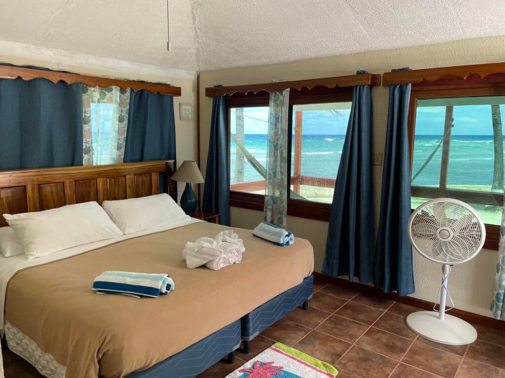 A cabana with a king bed with rolled towels on the bed, and windows showing views of the ocean beyond it's private terrace.
